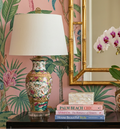 Decorative table setting with floral lamps, a mirror, and a plant against a pink wall with green leaf patterns.
