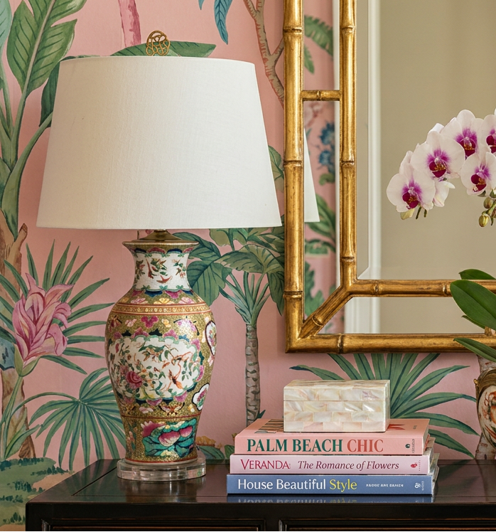 Decorative table setting with floral lamps, a mirror, and a plant against a pink wall with green leaf patterns.