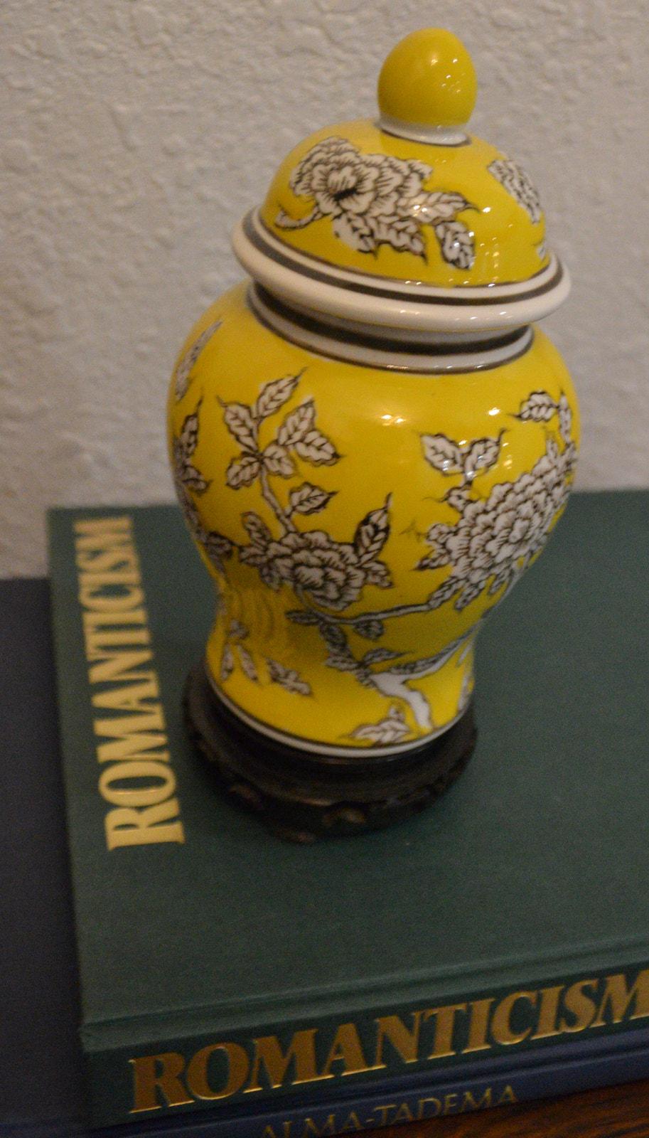 May include: A yellow ceramic jar with a lid, decorated with black and white floral patterns. The jar sits on a dark wooden base, and is placed on top of a green book titled 'Romanticism' with gold lettering. The jar has a rounded body and a small, round knob on the lid.